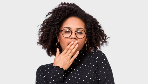 Giovane donna afroamericana stanca che sbadiglia, indossa camicetta nera e occhiali rotondi, posa sul muro bianco dello studio. Donna afro assonnata con capelli ricci disordinati, sentirsi stanchi, che copre la bocca aperta con il palmo della mano — Foto Stock