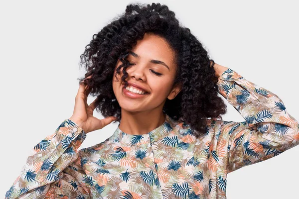 Freut sich eine dunkelhäutige junge Frau in floralem Hemd, die Hände auf dem Kopf haltend, glücklich. Afroamerikanerin lächelt breit und posiert über weiße Wand — Stockfoto