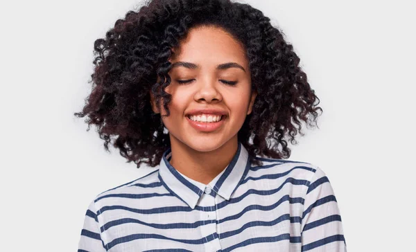 Chica de piel oscura soñadora feliz sonriendo ampliamente con los ojos cerrados, usando camisa a rayas, disfrutar del buen tiempo, posando sobre fondo blanco estudio. Gente, éxito, emociones y concepto de felicidad — Foto de Stock