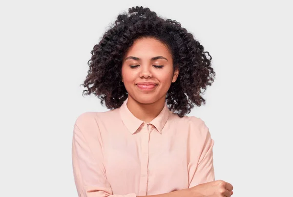 Vrolijke Afro-Amerikaanse jonge vrouw die lacht met gesloten ogen, het dragen van beige shirt met krullend afro haar, genieten van tijdig poseren over witte studio muur. Mensen, succes, emoties en geluk concept — Stockfoto