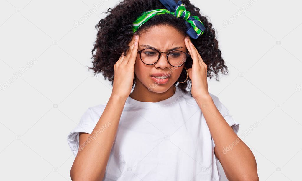 Exhausted African American young woman touching her head with closed eyes wearing white t-shirt,  green headband and eyewear. Afro female student suffering from unbearable headache. Health concept