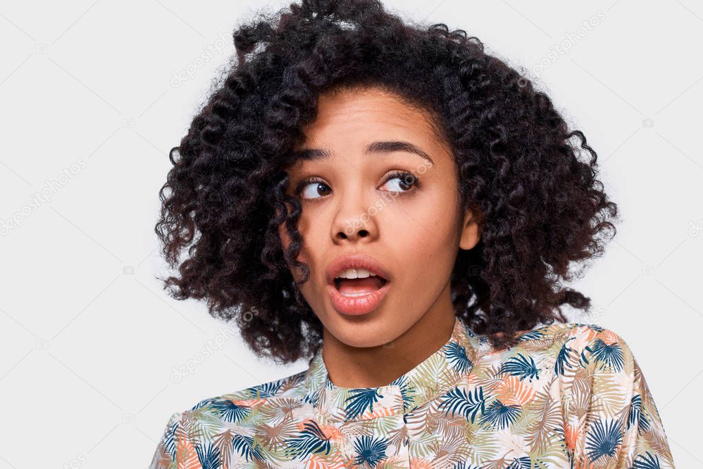 Surprised dark skinned yong woman wearing floral shirt, has astonished expression, looking at one side, posing over white wall background. African Americamn female has amazed expression.