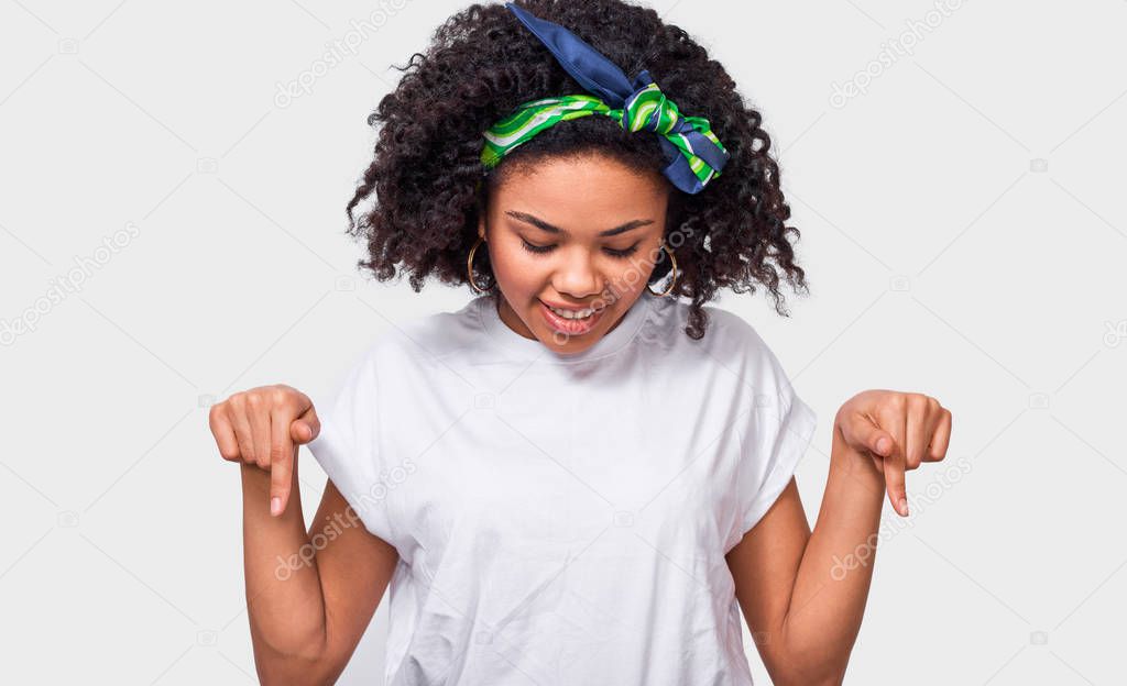 Studio shot of pretty young African American woman point at floor, wearing white t shirt, colorful headband, ont white wall. Happy dark skinned female indicates down with forefingers for advertising