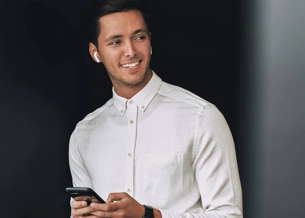 Happy young man smiling, texting messages from his cellphone over studio black background, with wireless earphones. Caucasian businessman wearing white shirt, using wireless earbuds for communication — Stock Photo, Image