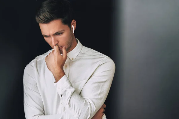 Portrait of sad young Caucasian handsome man speaking with a friend using wireless earphones, posing on black wall. Serious businessman using wireless earbuds during conversation. People, technology