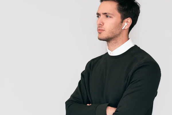 Serious young man standing with crossed hands and having a call via wireless earphones with a colleague isolated on white wall. Businessman using wireless earbuds for communication. Technology, people — Stock Photo, Image