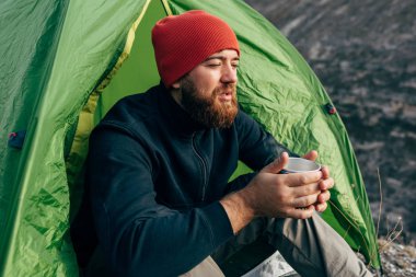 Dağlarda sıcak içecek içme, şenlik ateşi yakın oturan, trekking sonra rahatlatıcı genç explorer adam. Kırmızı şapkalı bir fincan çay sonra yürüyüş tutarak Gezgin adam. Seyahat, insanlar