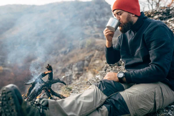 Bild eines jungen Entdeckers, der in den Bergen Heißgetränke trinkt, am Lagerfeuer sitzt und sich nach dem Trekking entspannt. Reisender Mann mit rotem Hut, der nach dem Wandern eine Tasse Tee in der Hand hält. Reisen, Menschen — Stockfoto