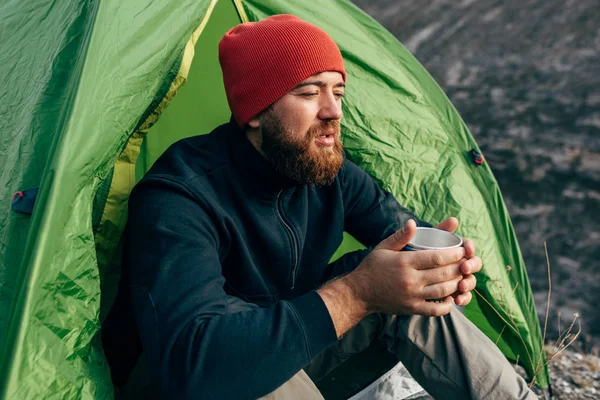 Junger Entdecker, der in den Bergen Heißgetränke trinkt, am Lagerfeuer sitzt und sich nach dem Trekking entspannt. Reisender Mann mit rotem Hut, der nach dem Wandern eine Tasse Tee in der Hand hält. Reisen, Menschen — Stockfoto