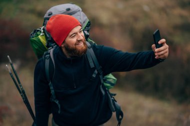 Gülümseyerek, akıllı telefonundan dağlarda selfie alarak mutlu genç adam. Gezgin sakallı erkek giyen kırmızı şapka yürüyüş sonra cep telefonundan kendi kendine portre almak. Seyahat, yaşam biçimi