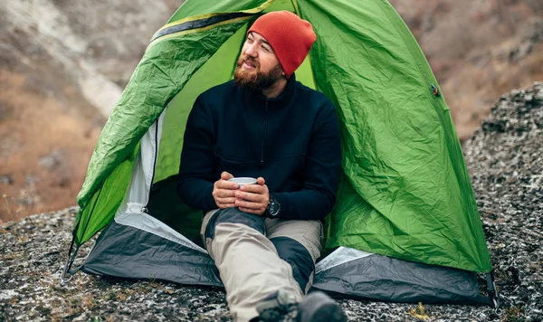 Horizontale Außenaufnahme eines bärtigen Mannes, der in der Nähe des Zeltes sitzt und sich nach dem Bergsteigen entspannt. Junge Wanderer genießen die Luft in den Bergen. Reisen, Menschen und gesunder Lebensstil — Stockfoto