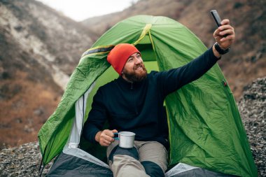 Gülümseyerek, akıllı telefonundan dağlarda selfie alarak mutlu genç adam açık havada vurdu. Gezgin kırmızı şapka giyen erkek portresi cep telefonu üzerinden yürüyüş sonra al. Seyahat, yaşam, teknoloji