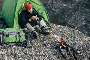 Havai otdoors atış bir şenlik ateşi yakın oturan dağlarda sıcak içecek içme genç explorer adam trekking sonra rahatlatıcı. Kırmızı şapkalı bir fincan çay sonra yürüyüş tutarak Gezgin adam. Seyahat