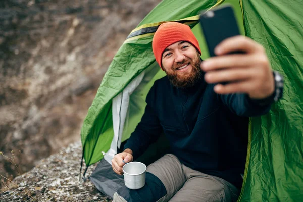 Imagem de um jovem feliz a sorrir, tirando selfie nas montanhas do seu telemóvel inteligente. Homem barbudo viajante usando chapéu vermelho tirar auto retrato do celular depois de caminhar. Viagens, estilo de vida, tecnologia — Fotografia de Stock