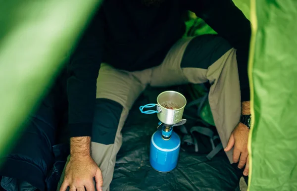 Abgeschnittener Kopfschuss eines jungen Entdeckers, der in den Bergen heiße Getränke zubereitet. Ein bärtiger Mann sitzt im Zelt und bereitet sich nach der Wanderung einen Becher Tee zu. Reisen, Menschen und helathischer Lebensstil — Stockfoto