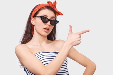 Görüntü trendy güneş gözlüklü genç esmer kadın hayran, kenara boş kopya alanı, beyaz duvardan izole için işaret parmağı ile gösterir. Açık ağız ile çizgili bluz giyen beyaz kız.