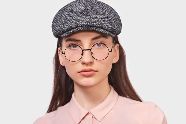Närbild studio porträtt av ganska ung kvinna bär transparenta glasögon, casual rosa skjorta och trendiga grå mössa, ser allvarligt direkt i kameran, poserar över vita studio vägg. Människor känslor — Stockfoto