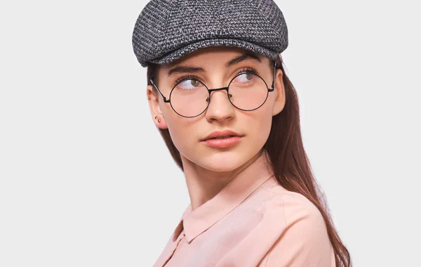 Närbild porträtt av vacker ung kvinna bär transparenta glasögon, casual rosa skjorta och trendiga grå mössa, ser allvarligt undan till kampanjtext, poserar över vita studio vägg. Människor känslor — Stockfoto
