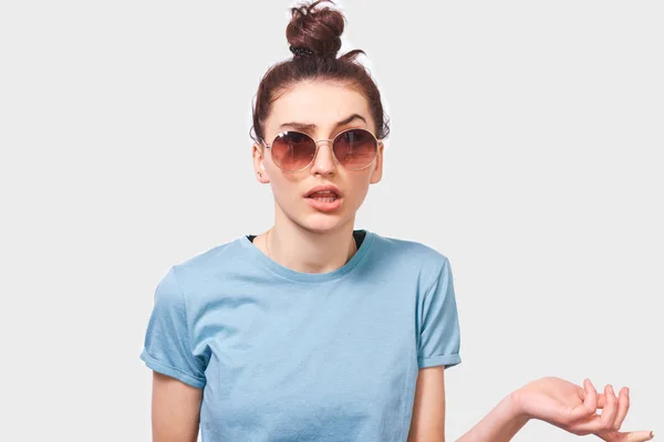 Mujer joven interrogada, mirando en serio a través de gafas de sol durante la discusión, vestida con camiseta azul con moño de pelo, posando sobre la pared blanca. La estudiante tiene expresión confusa. Personas emociones —  Fotos de Stock