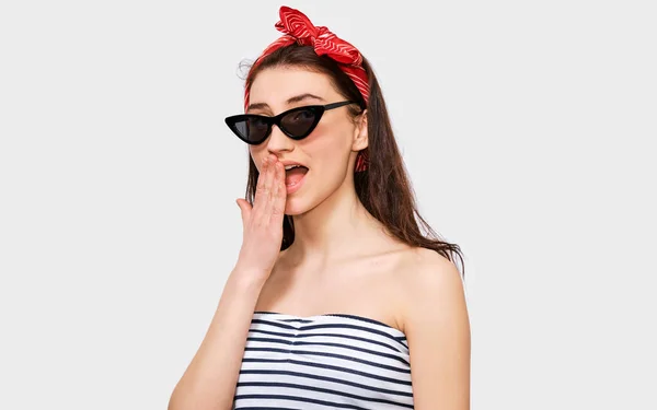Upphetsad vacker ung kvinna bär randig blus, svart trendiga solglasögon och rött pannband, ser att kamera, isolerade på vita studio bakgrund. Flicka med handen på munnen har förvånat uttryck. — Stockfoto