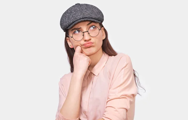 Pensive young woman wears transparent spectacles, casual pink shirt and gray cap, looks up to the promotional text, poses against white studio background. Serious girl thinking about an idea. People — Stock Photo, Image