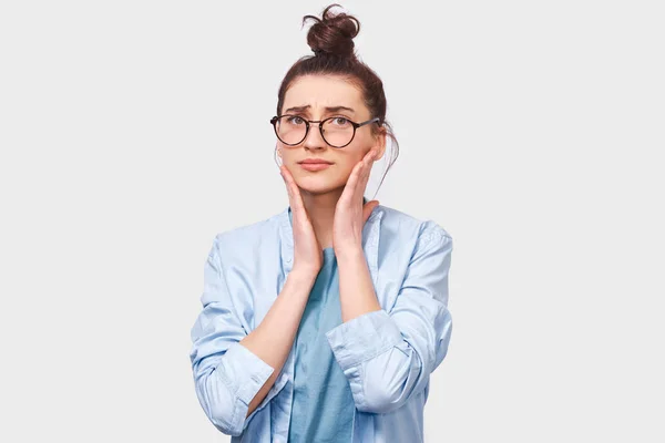 Mujer joven preocupada, mirando directamente a la cámara a través de gafas redondas. Chica dudosa lleva camisa azul no puede hacer elección, ha cuestionado la expresión con las manos en la cara, posando sobre la pared blanca . —  Fotos de Stock