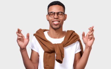 Arzulu genç Afro-Amerikan adam diş clenches, parmak çarpı işareti yükseltir, arzu dilek yapar, Beyaz tişört, beyaz duvara poz giyer. Karışık yarış erkek parmak sınavlarına haçlar. İnsanlar, duygular