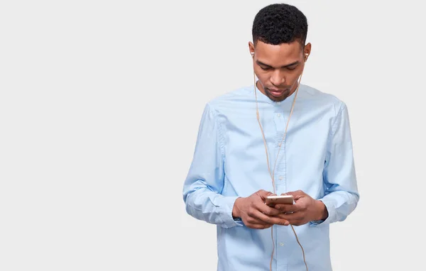 Горизонтальный студийный портрет афроамериканского молодого человека, который пишет сообщения и ведет блог по мобильному телефону. Красивый мужчина слушает музыку на наушниках через мобильный телефон, с пустым местом для копирования. Человек, технологии — стоковое фото