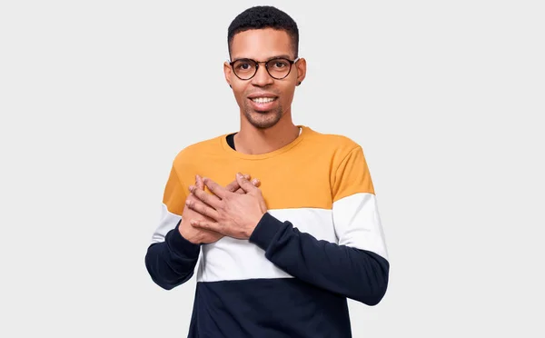 Candid portrait of kind dark-skinned man keeps both palms on chest, expresses kindness. Afro male looks directly to camera, in casual outfit, isolated over white background. People and body language — Stock Photo, Image