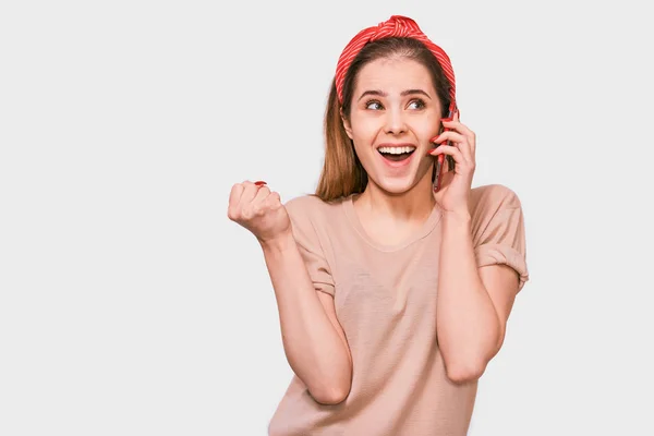 Mulher bonita nova em camiseta bege, headband vermelho, sorrindo e falando no telefone inteligente para sua amiga, olhando alegre e feliz, posando no fundo do estúdio branco. Emoções humanas reais . — Fotografia de Stock