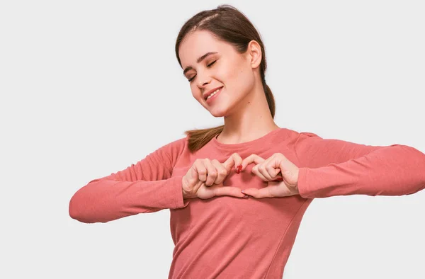 Smiling young woman making heart shape with fingers. Beautiful female with closed eyes, dressed in pink blouse, showing heart gesture with two hands, love sign, posing on white background. — Stock Photo, Image
