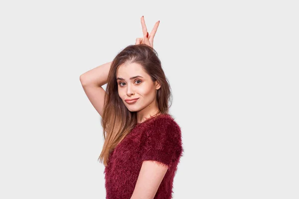 Funny Caucasian young woman smiling and showing peace gesture while looking to the camera and standing against white background. Happy female shows her emotions, making ears with two fingers. — Stock Photo, Image