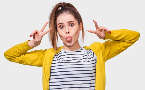 Portrait of funny young woman showing tongue and peace gesture while looking to the camera and standing against white background. Happy female wearing casual outfit shows her emotions. — Stock Photo, Image