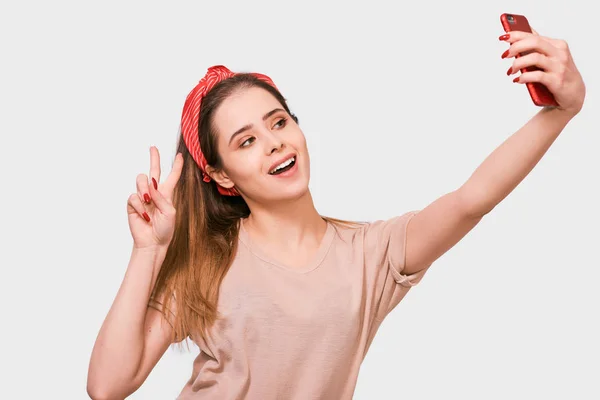 Beautiful young European woman in casual clothes and red headband, taking self portrait over white studio wall. Happy female smiling and taking a selfie on her smart phone. — Stock Photo, Image