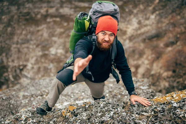 Horizontal image of traveler bearded man trekking and mountaineering during his journey. Young tourist male holds out his hand for help during hiking in mountains. Travel, people, sport, lifestyle
