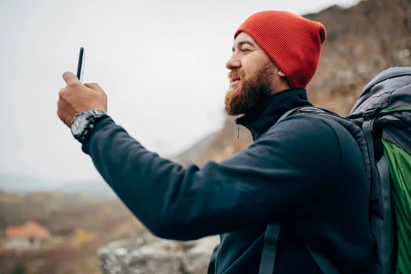 Portrait latéral d'un jeune homme prenant des photos en montagne depuis son téléphone intelligent. Voyageur barbu mâle portant chapeau rouge prendre selfie de téléphone mobile après la randonnée. Voyages, mode de vie — Photo