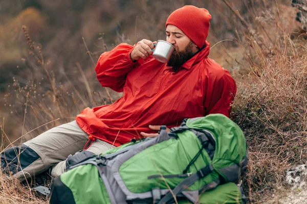 Young explorer man drinking hot beverage in mountains, sitting and relaxing after trekking. Traveler man in red hat holding a mug of tea after hiking. Travel, people — Stock Photo, Image