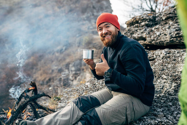 Image of happy young explorer man drinking hot beverage in mountains, sitting near to bonfire, relaxing after trekking. Traveler man in red hat holding a mug of tea after hiking. Travel, people