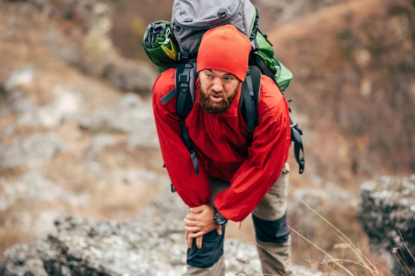 Image of tired hiker man hiking in mountains dressed in red clothes exploring new places. Traveler bearded male trekking and mountaineering during his journey. Travel, people, sport, lifestyle concept