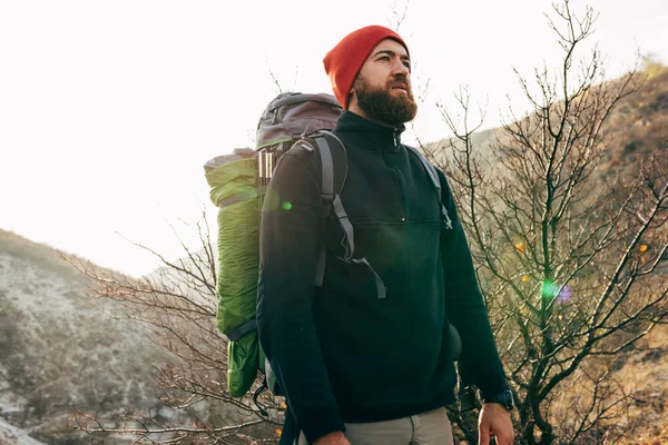 Utendørs portrett av en ung mann med utsikt mot horisonten i fjell med reiseryggsekk og rød hatt. Reisende skjeggete mann som slappet av etter å ha gått tur under reisen. Reise, livsstil, sport – stockfoto