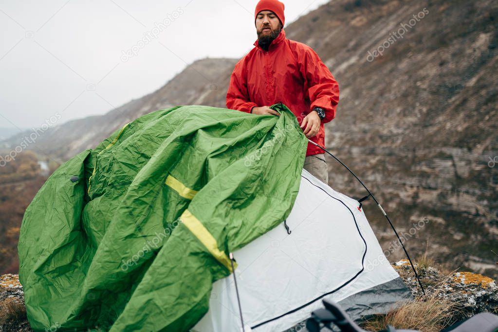 Tourist young hiker man prepares his tent in mountains. Traveler bearded male relaxing after mountaineering. Travel, people and healthy lifestyle concept