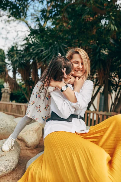 Ładne córka obejmując i Całowanie jej młody szczęśliwy matka ubrany w żółty spódnica siedzący w The Park. Piękna kobieta przytulanie dziecka dziecko na zewnątrz. Dzień matki. Szczęśliwa rodzinna relacja — Zdjęcie stockowe