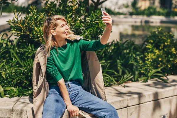 Pen jente som lager selvportrett med rar grimase. Ung, glad kvinne smiler bredt mens hun holder smarttelefonen gående utendørs i byen på solfylte dager. Lykkelig kvinne som tar selfie på mobiltelefon . stockbilde