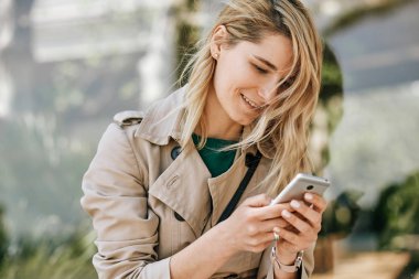 Genç güzel kadın gülümseyen ve akıllı telefon mesajları yazma Closeup portre, güneşli bir günde şehirde açık oturan. Güzel kadın cep telefonundan haber okuyor. Seyahat ve teknoloji konsepti