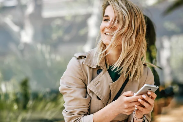 Retrato de jovem feliz bela mulher sorrindo e olhando para longe, usando telefone celular, sentado ao ar livre na cidade no dia ensolarado. Uma mulher bonita a ler notícias no telemóvel. Conceito de viagem e tecnologia — Fotografia de Stock