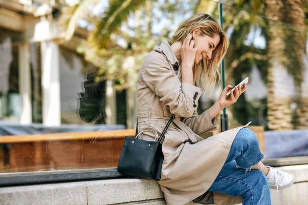 Горизонтальное изображение молодой красивой женщины, пишущей сообщения на смартфоне, сидящей на открытом воздухе в городе в солнечный день. Красивая женщина с бесплатным беспроводным телефоном. Концепция путешествий и технологий — стоковое фото