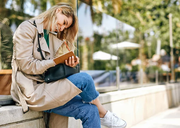 Jovem alegre e franca sorrindo amplamente enquanto conversa com um amigo no smartphone, sentada ao ar livre na cidade no dia ensolarado. Mulher bonita falando no celular, escrevendo alguns avisos no caderno . — Fotografia de Stock