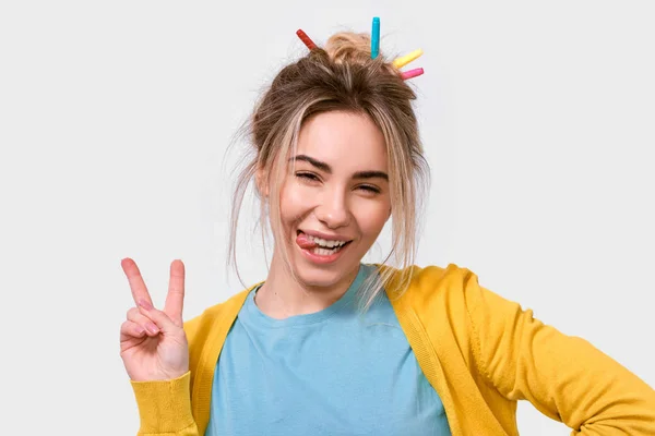 Jovem mulher alegre mostrando língua e gesto de paz enquanto olha para a câmera e de pé contra o fundo branco. Mulher feliz vestindo roupas casuais e marcadores coloridos no cabelo se divertindo — Fotografia de Stock