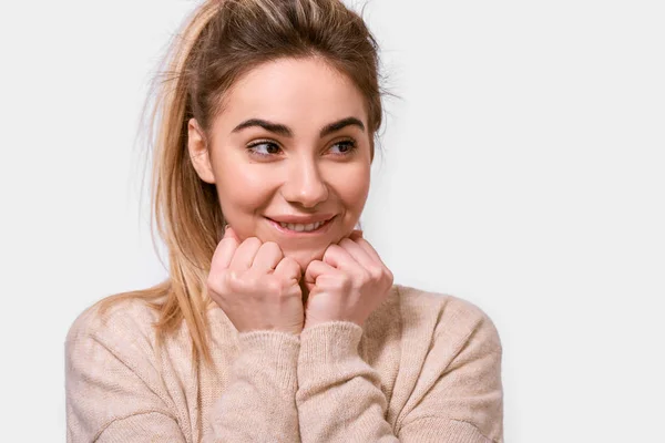Close up portrait of cute kind young woman smiling and keeps hands on chin, looking aside, dressed beige sweater, isolated over white background. People, emotions, feeling and body language concept — Stock Photo, Image