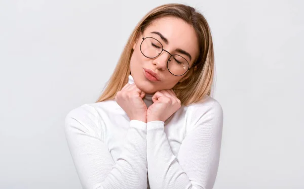 Beautiful young woman keeps hands on chest with closed eyes, wearing white blouse and round eyeglasses, isolated over white background. People, emotions, feeling and body language concept — Stock Photo, Image